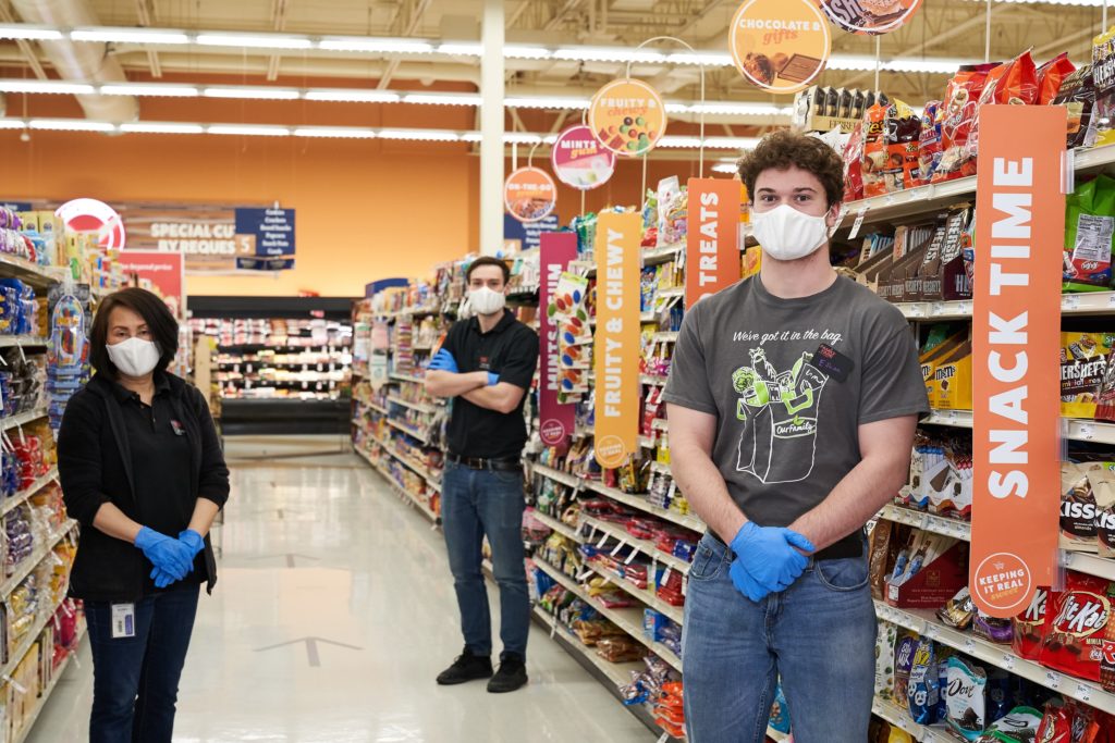 Grocery store workers wearing pandemic masks and rubber gloves, standing socially distanced in aisle