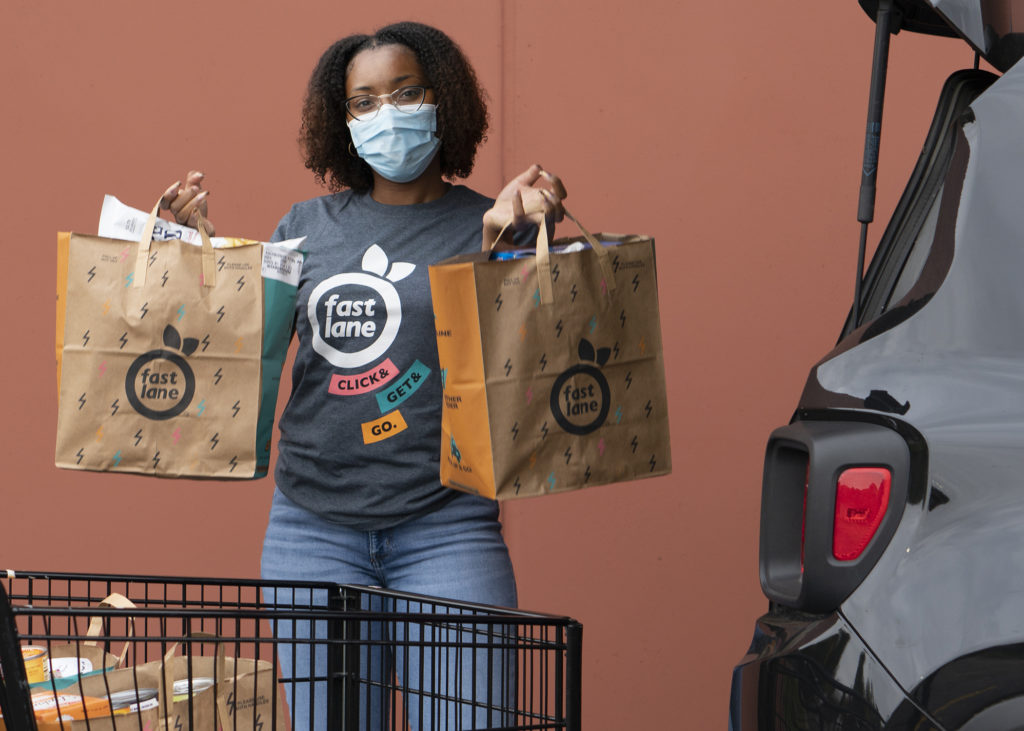 African American woman in medical mask holding groceries in paper bags by the hatch of a compact SUV
